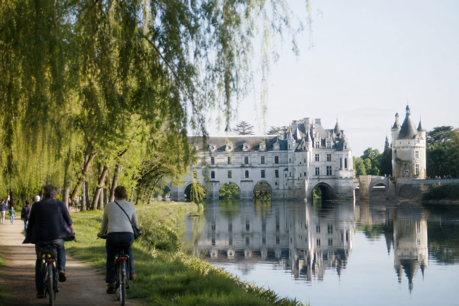 Château de tuffeau blanc reflété dans une rivière calme, cyclistes sur le chemin de halage
