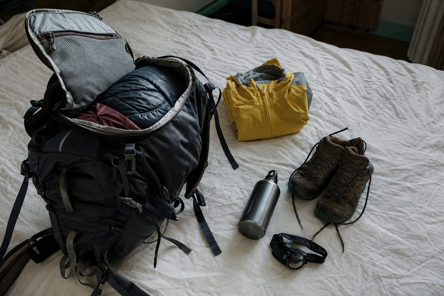 Sac à dos ouvert sur un lit avec vêtements de pluie et chaussures de marche