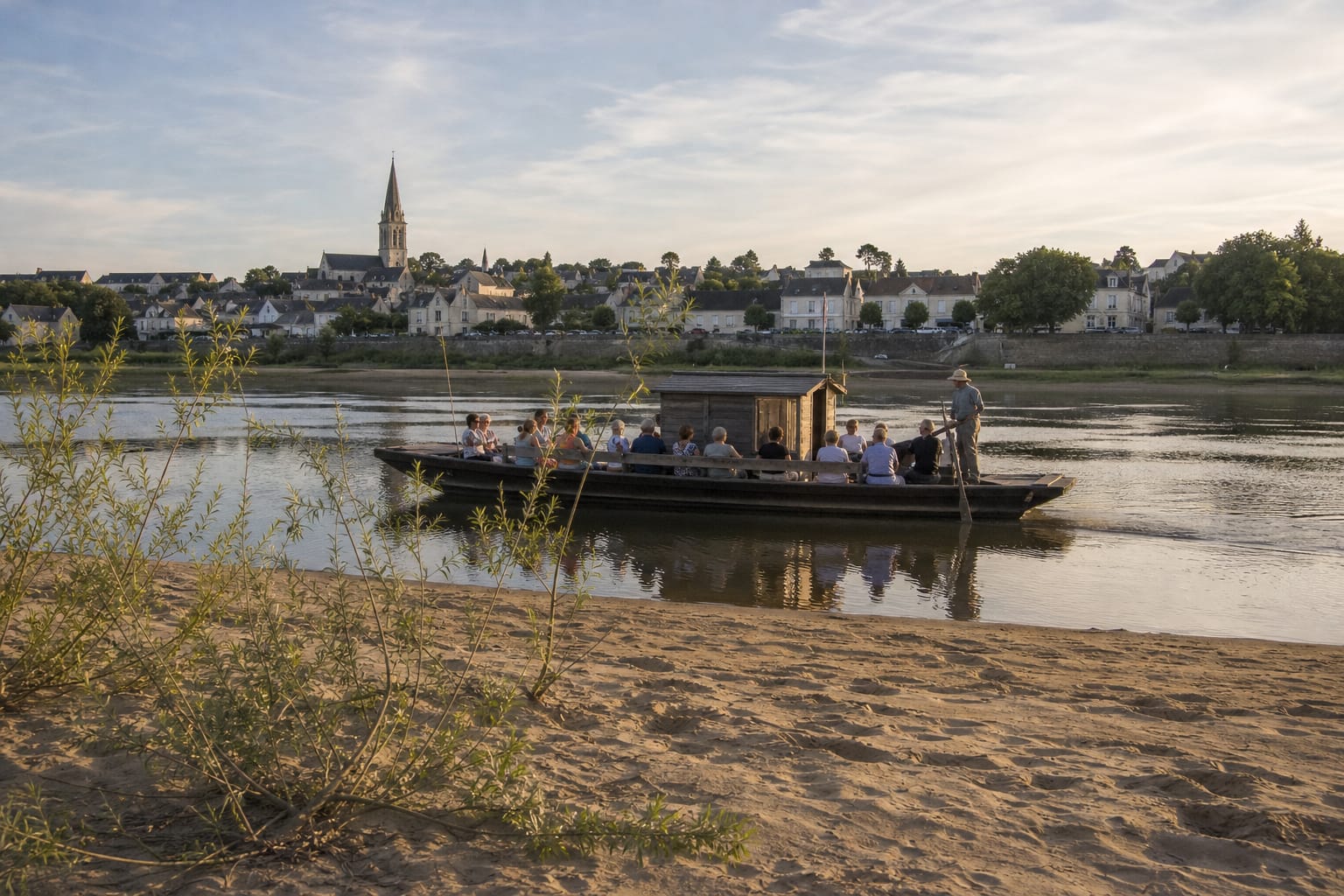 Toue traditionnelle en bois chargée de passagers glissant sur la Loire en fin de journée, devant un village de tuffeau et un banc de sable bordé de saules