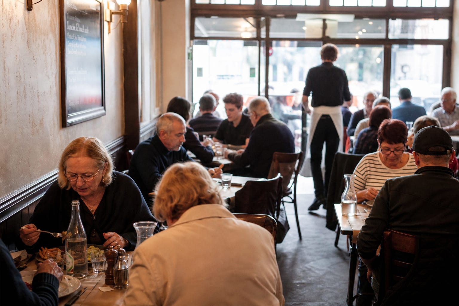 Salle de bistrot à midi, ardoise du jour accrochée au mur, tables occupées