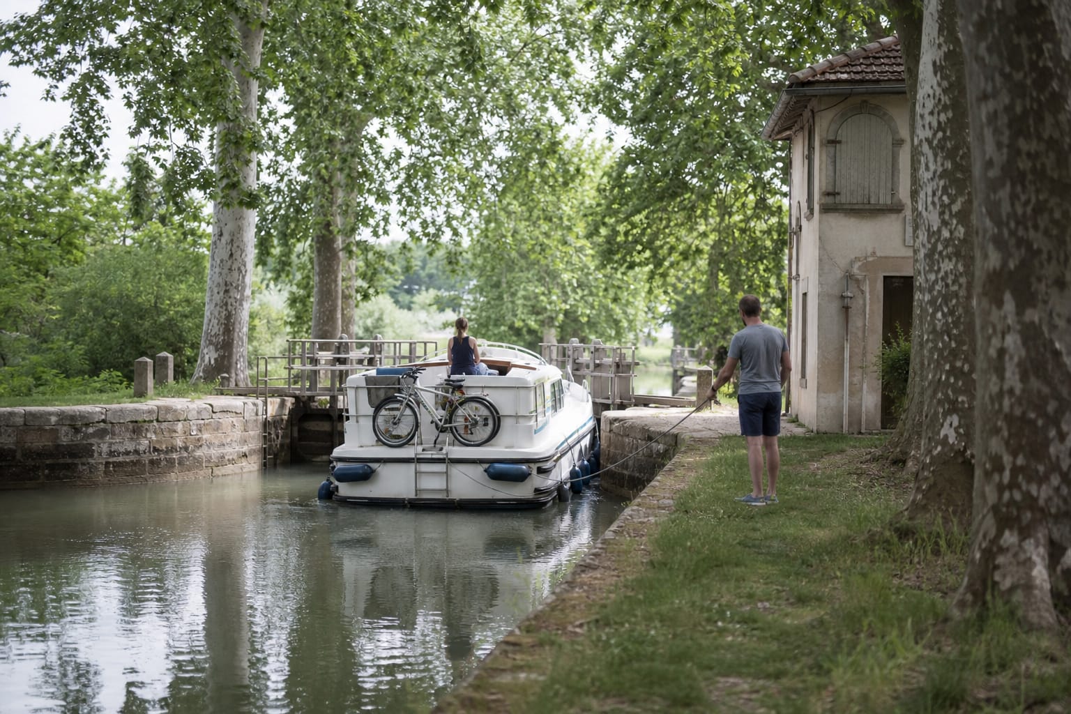 Bateau habitable sortant d'une écluse sur un canal bordé de platanes, un équipier tenant l'amarre sur la berge en fin de matinée